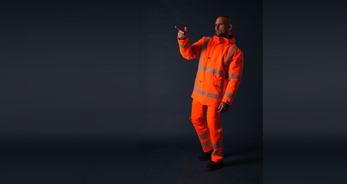 Man in fluororanje hi-vis jas met bijpassende broek, met reflecterende banden om romp, mouwen en broekspijpen.