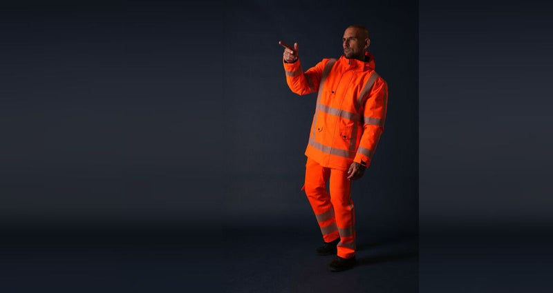Man in fluororanje hi-vis jas met bijpassende broek, met reflecterende banden om romp, mouwen en broekspijpen.